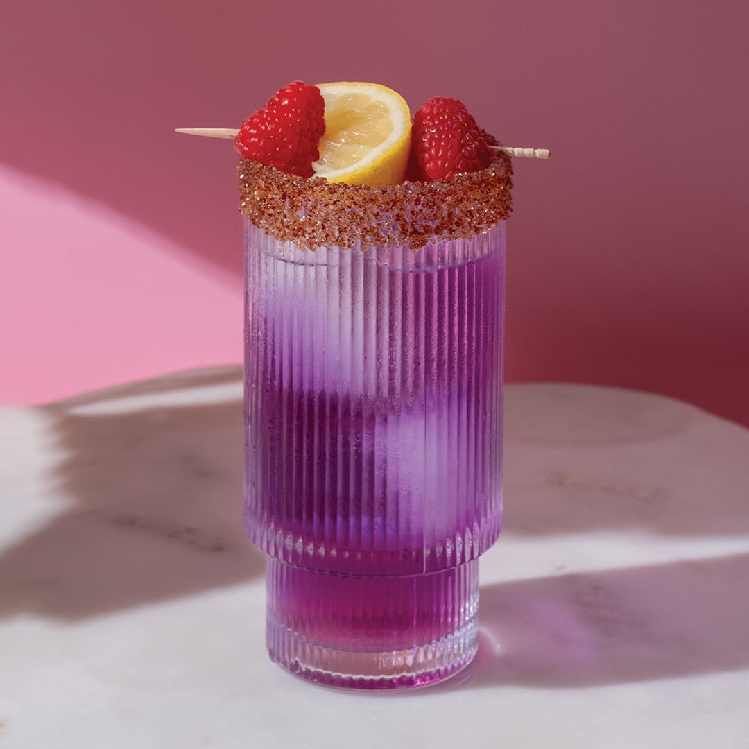 A tall, ribbed glass filled with a purple cocktail and ice sits on a white surface. The glass has a sugared rim and is garnished with a lemon slice and two raspberries on a toothpick. The background is pink.